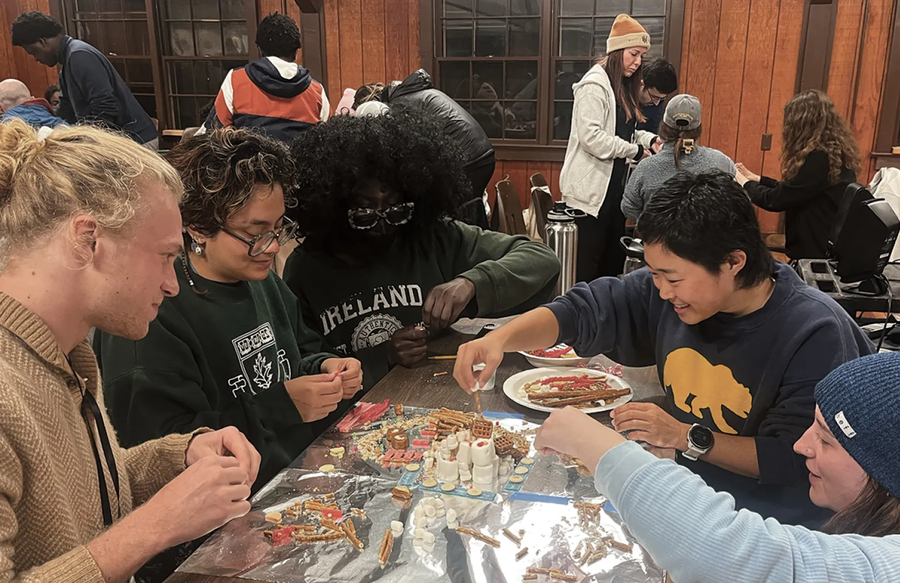 A group of young adults sit around a table indoors making a craft project with pretzels, marshmallows, and candy, smiling and concentrating as they work.