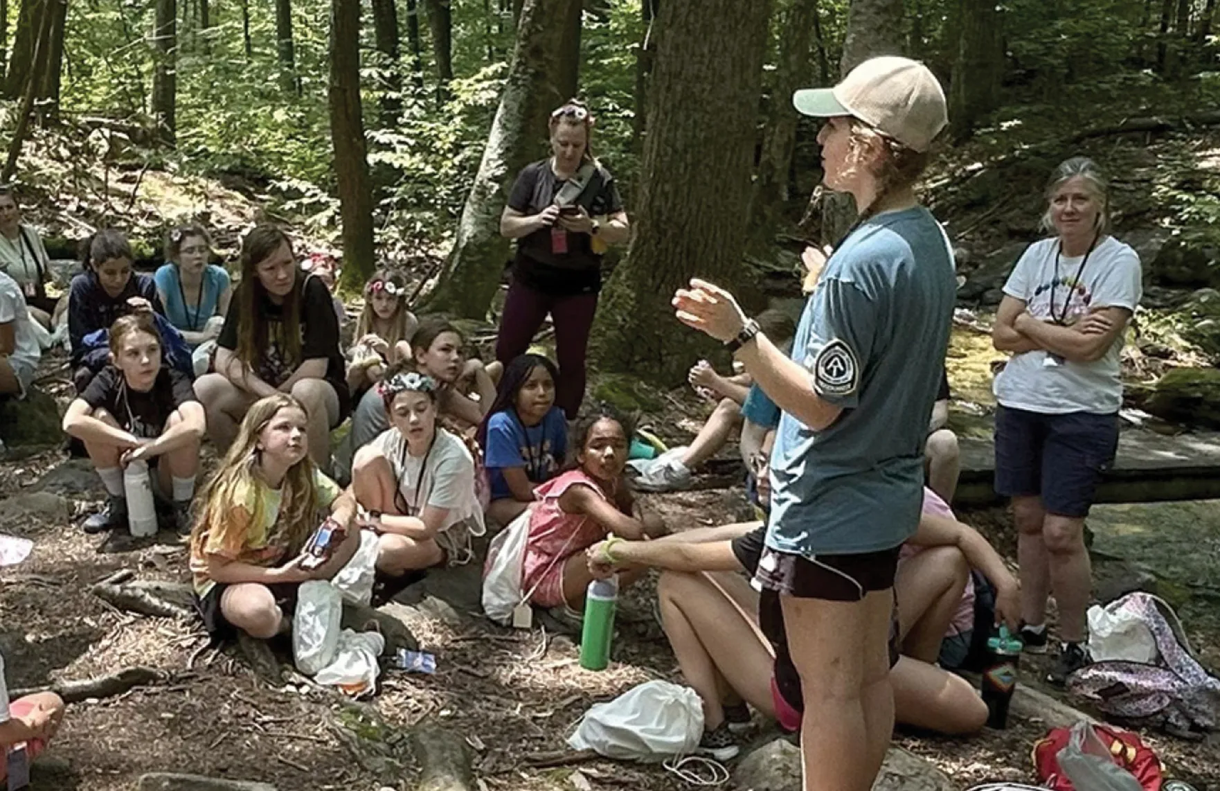 A woman in an Appalachian Trail shirt speaks to a group of children seated on the forest floor during an outdoor educational lesson, with trees surrounding them.