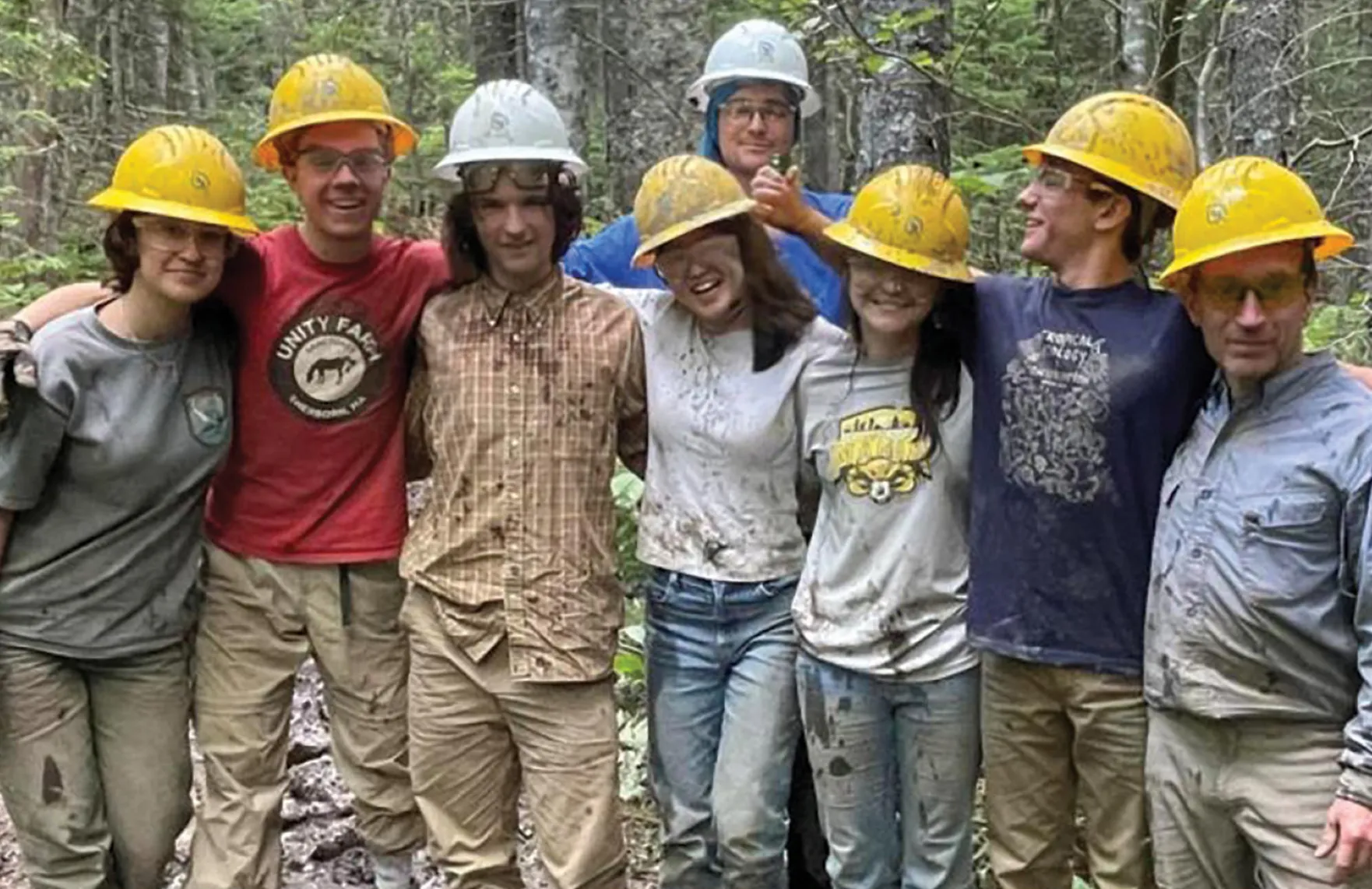 A group of trail crew members wearing hard hats and safety glasses stand arm in arm in a forest, covered in mud after a day of trail work.