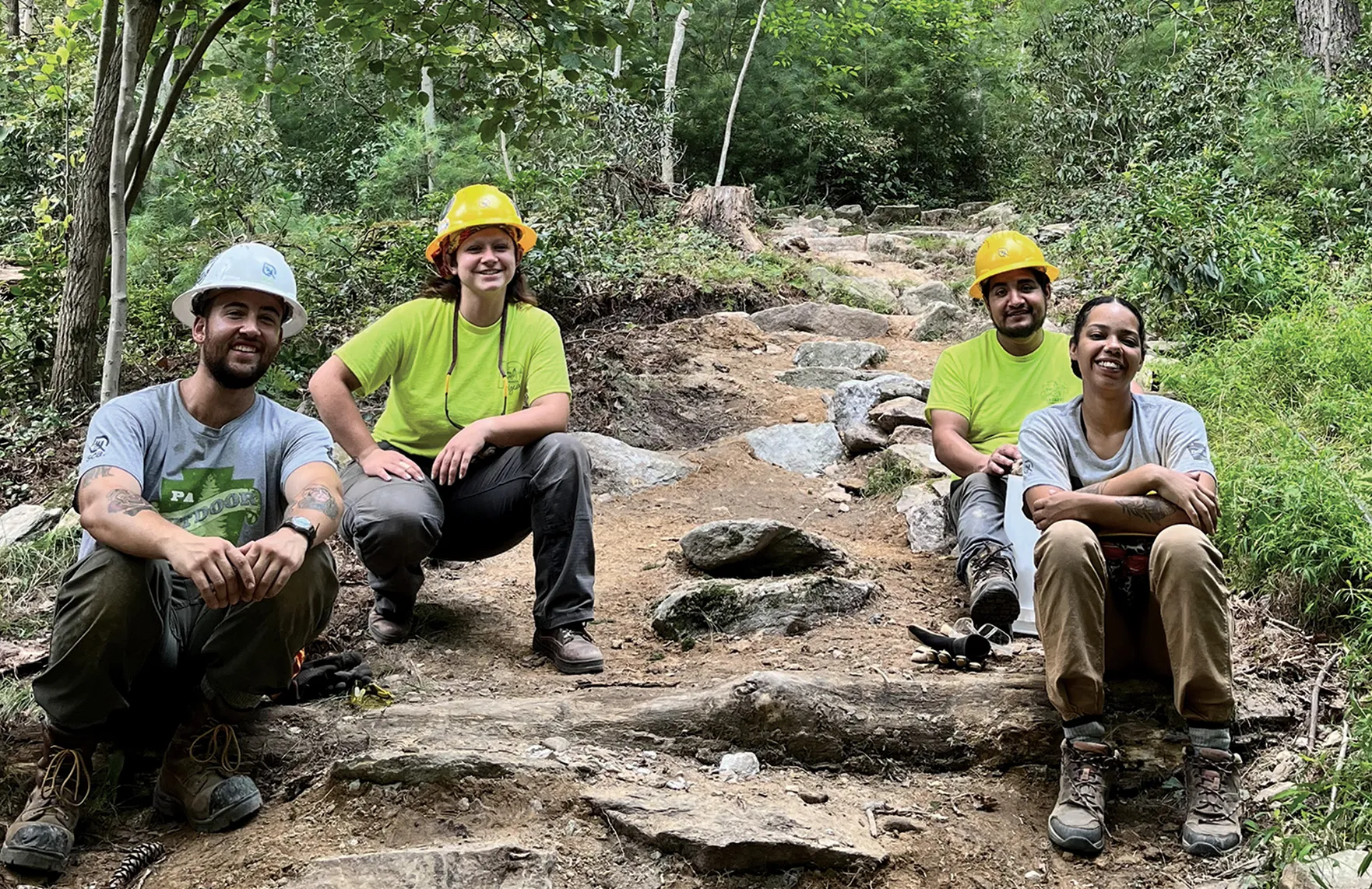 Four trail workers wearing hard hats rest on a rocky section of trail they are building or repairing, smiling and sitting among stones and tree roots.
