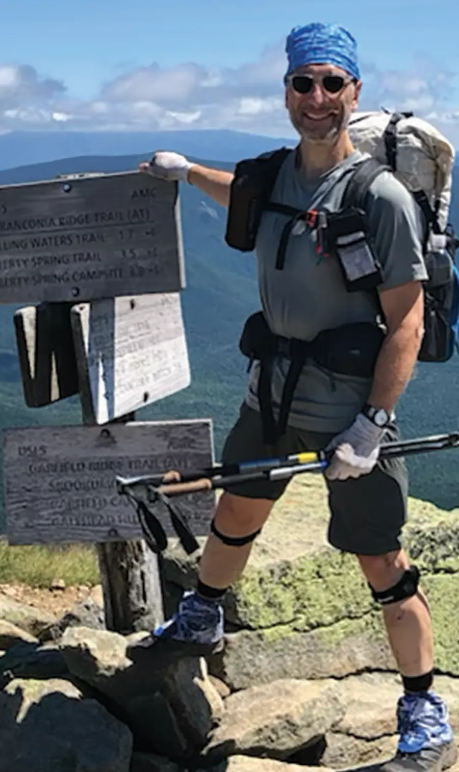 Jim LaTorre, a smiling hiker man wearing sunglasses and a blue bandana, stands on a rocky mountain summit, holding hiking poles; He's wearing a backpack and shorts; Beside him is a wooden trail marker sign with text like FRANCONIA RIDGE TRAIL and distances listed for various locations; The background features a sweeping view of green, mountainous terrain under a blue sky with some clouds