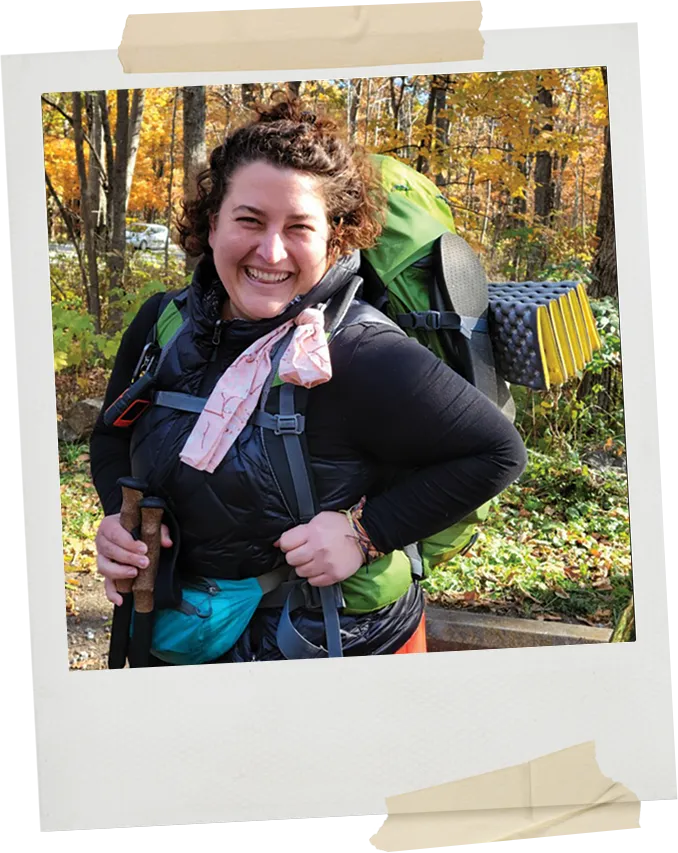 A smiling hiker wearing a large backpack and holding trekking poles poses in a forest during autumn, surrounded by colorful fall foliage.