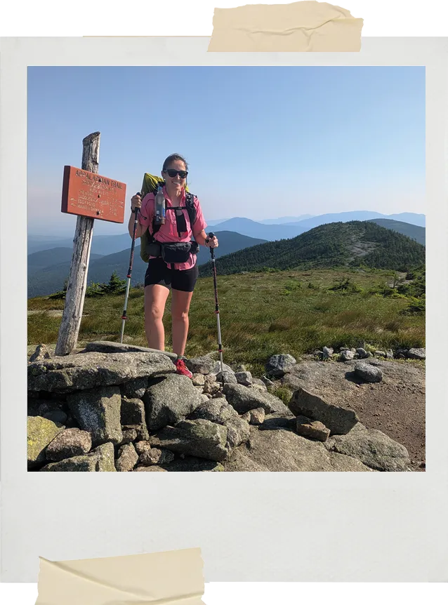 A hiker in a pink shirt and backpack stands beside an Appalachian Trail sign on a rocky summit with distant mountains visible behind her.