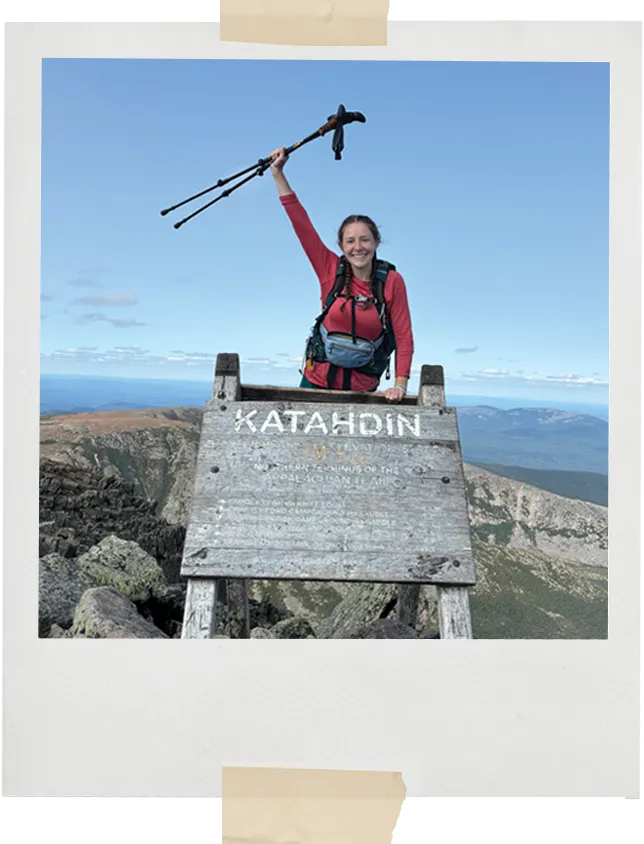 A hiker raises trekking poles triumphantly while standing at the wooden Katahdin summit sign with rugged mountain scenery in the distance.