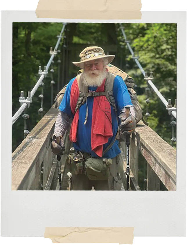 An older bearded hiker wearing a sun hat and blue shirt walks across a suspension bridge with trekking poles, surrounded by lush green forest.