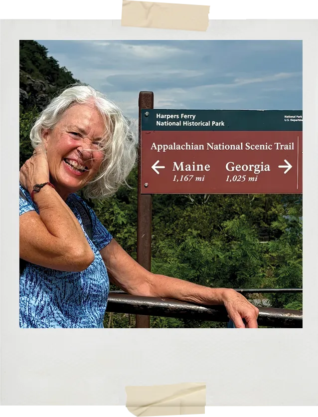 A smiling woman poses beside a sign at Harpers Ferry indicating the Appalachian Trail distances to Maine and Georgia, with trees and sky in the background.