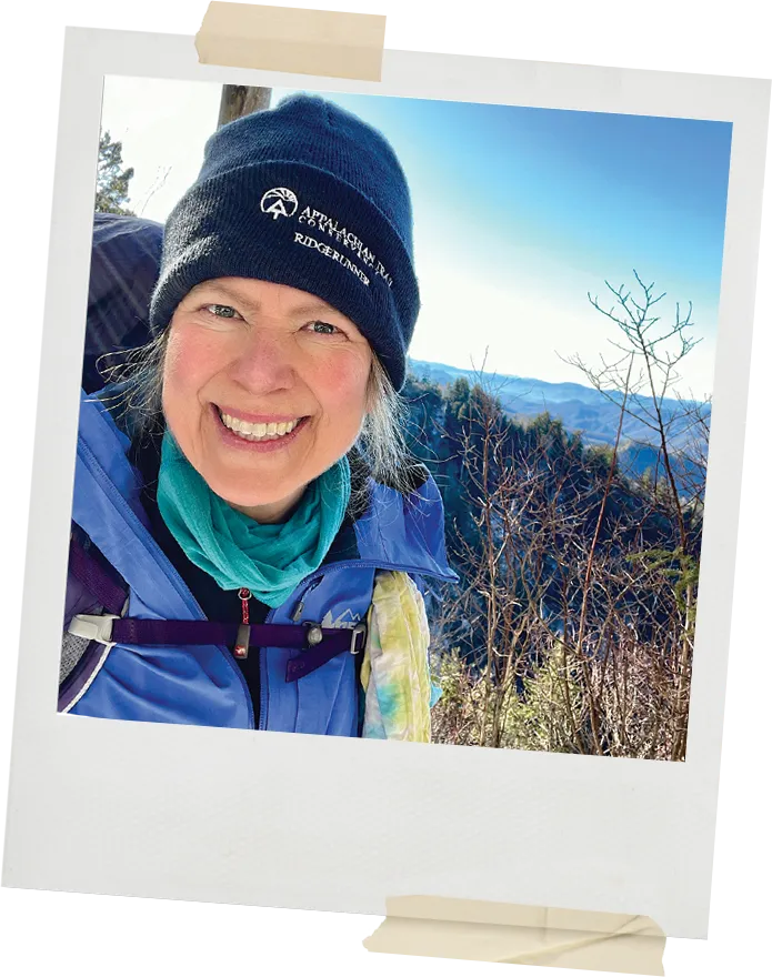 A woman wearing an Appalachian Trail Conservancy hat smiles outdoors with mountain ridges and winter trees behind her.