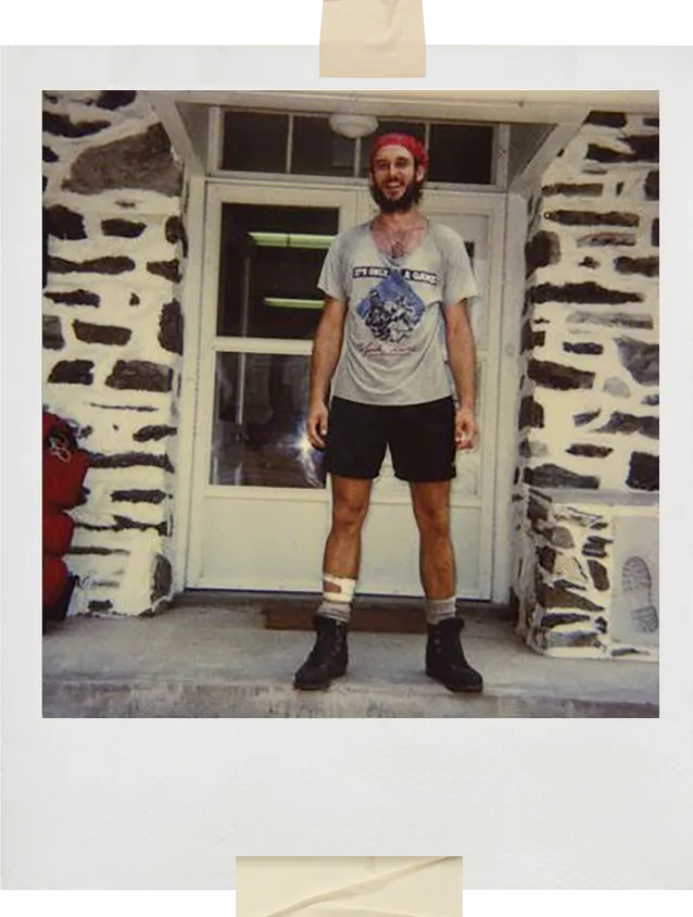 A vintage Polaroid-style photo of a smiling hiker standing in front of a stone building. He wears a red bandana, a gray graphic T-shirt, dark shorts, thick socks, and worn hiking boots. A red pack is visible to the left of the doorway behind him.