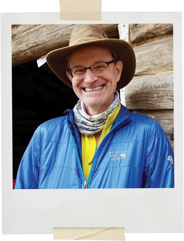 A Polaroid-style portrait of a smiling man in a brown brimmed hat, glasses, and a bright blue Mountain Hardwear jacket layered over a yellow shirt. He stands in front of a rustic wooden cabin wall.