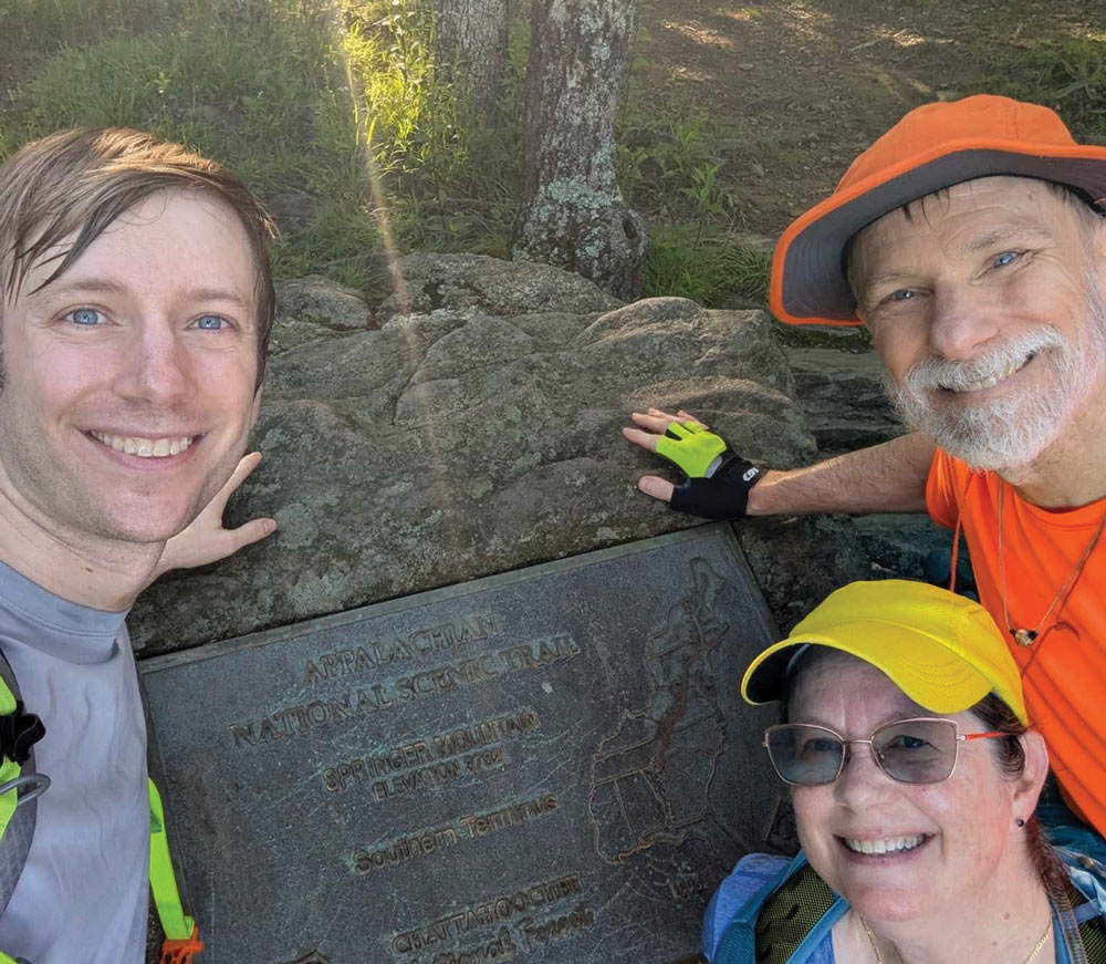 Three hikers smiling and touching the Appalachian Trail Southern Terminus plaque on Springer Mountain.