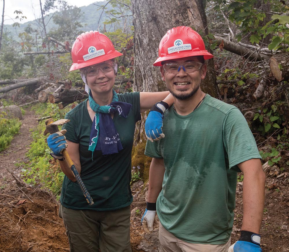 A man and woman wearing red hard hats and safety glasses hold trail maintenance tools in a wooded area.