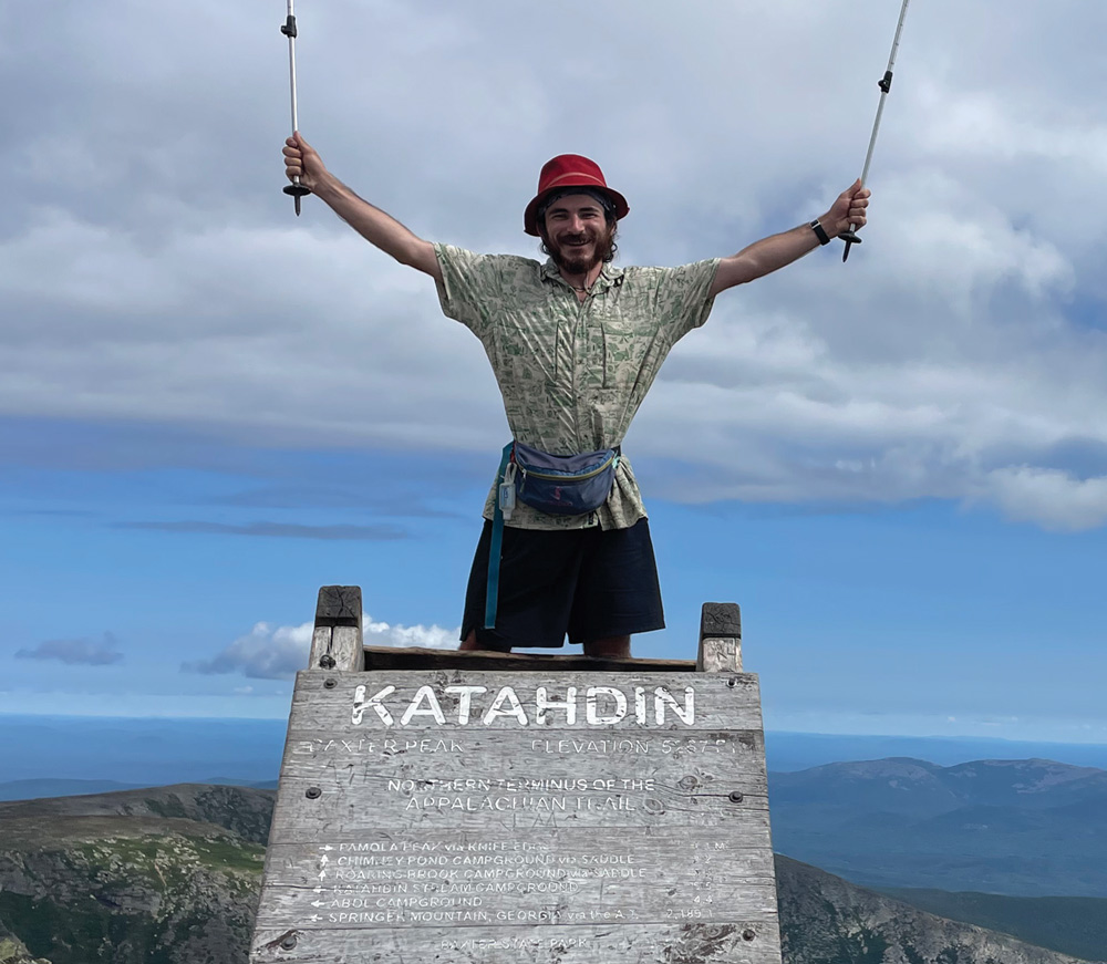 A hiker wearing a red bucket hat stands on the Katahdin summit sign with trekking poles raised in triumph.