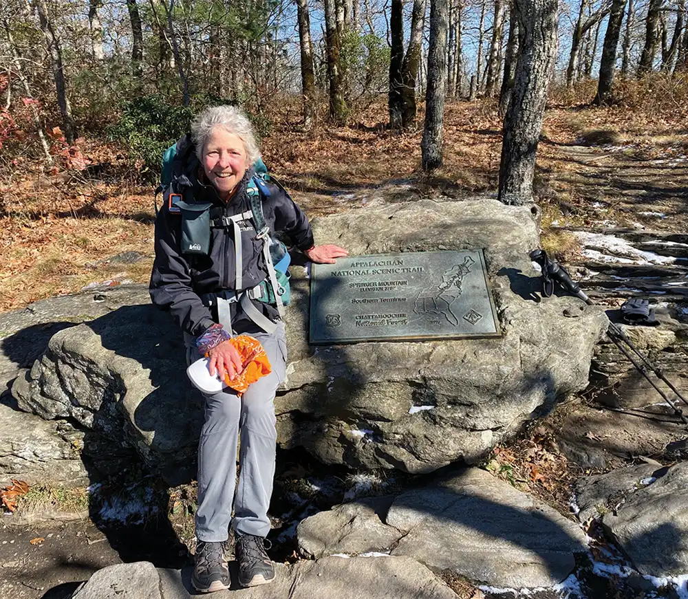 A female hiker with gray hair and a backpack sits on a rock next to the Appalachian Trail plaque at Springer Mountain.