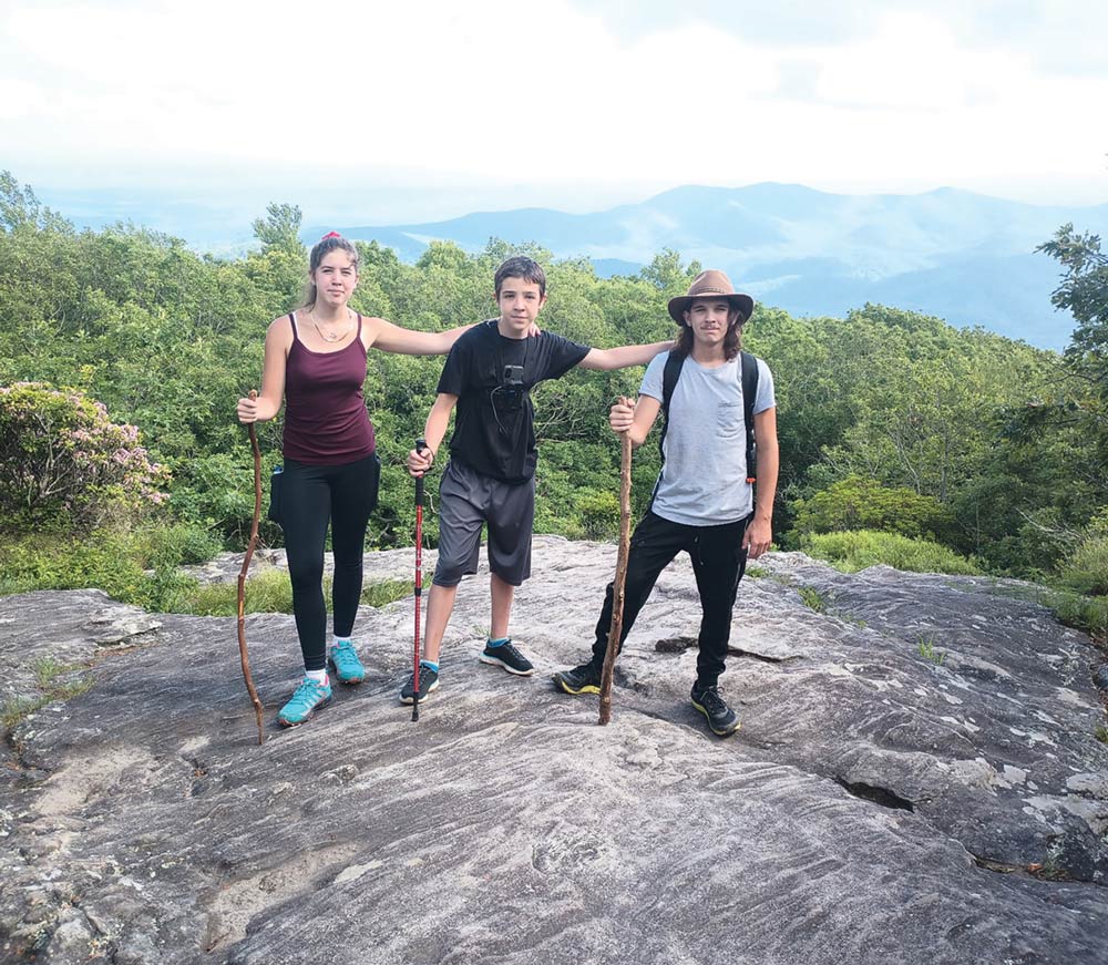 Two young men and a young woman pose on a rocky mountain peak, holding wooden walking sticks with a mountain view.
