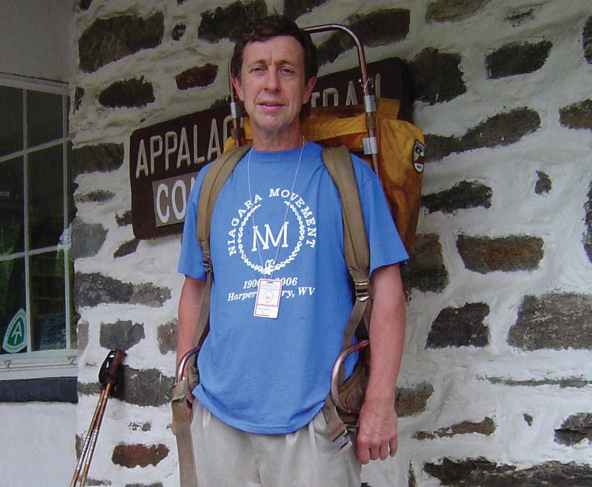 A man with a vintage external frame backpack stands in front of a stone building with an "Appalachian Trail Conservancy" sign.