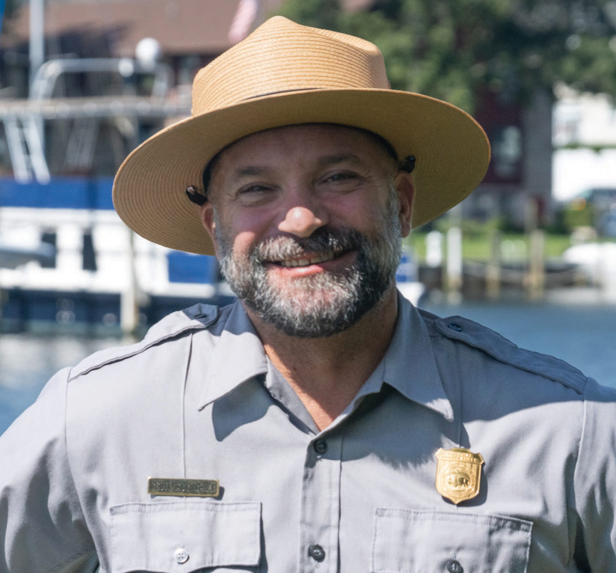 A close-up portrait of a smiling man wearing a tan wide-brimmed ranger hat and a grey uniform with a gold badge.