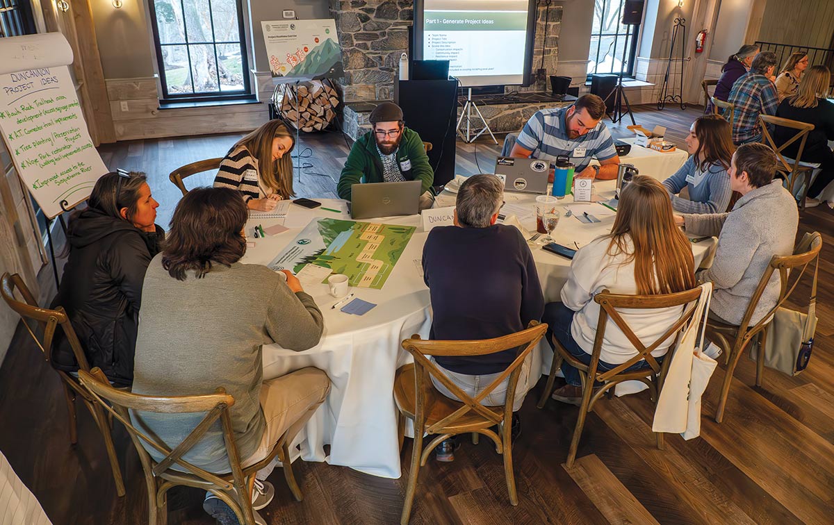 A group of people sitting at a round table during a community workshop, using laptops and reviewing project posters.