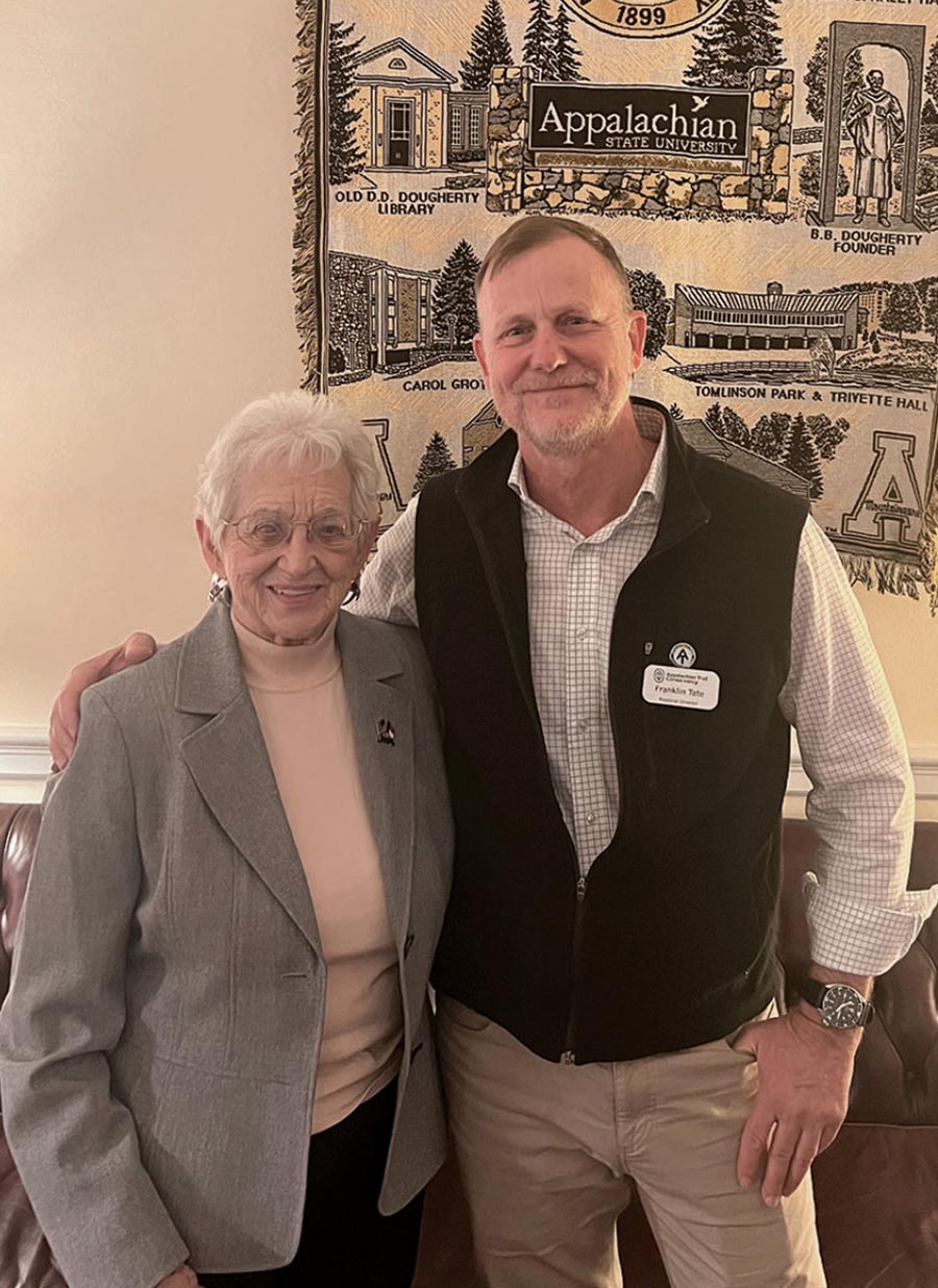 An older woman in a grey blazer stands next to a smiling man in a black fleece vest. Behind them hangs a large tapestry with illustrations of Appalachian State University campus buildings.