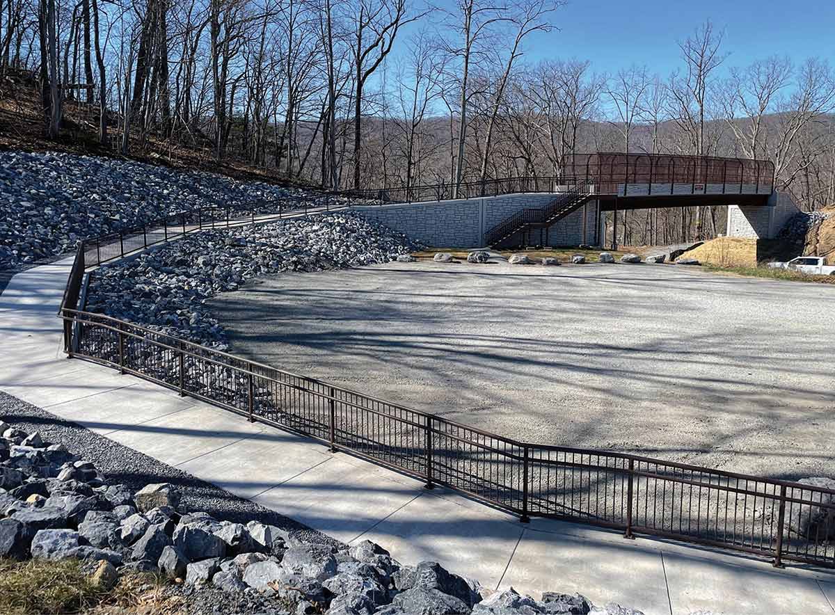 Paved walkway with metal railings curving past a rocky slope and gravel area toward a pedestrian bridge with stone walls. Bare trees and mountains are in the background under a clear sky.