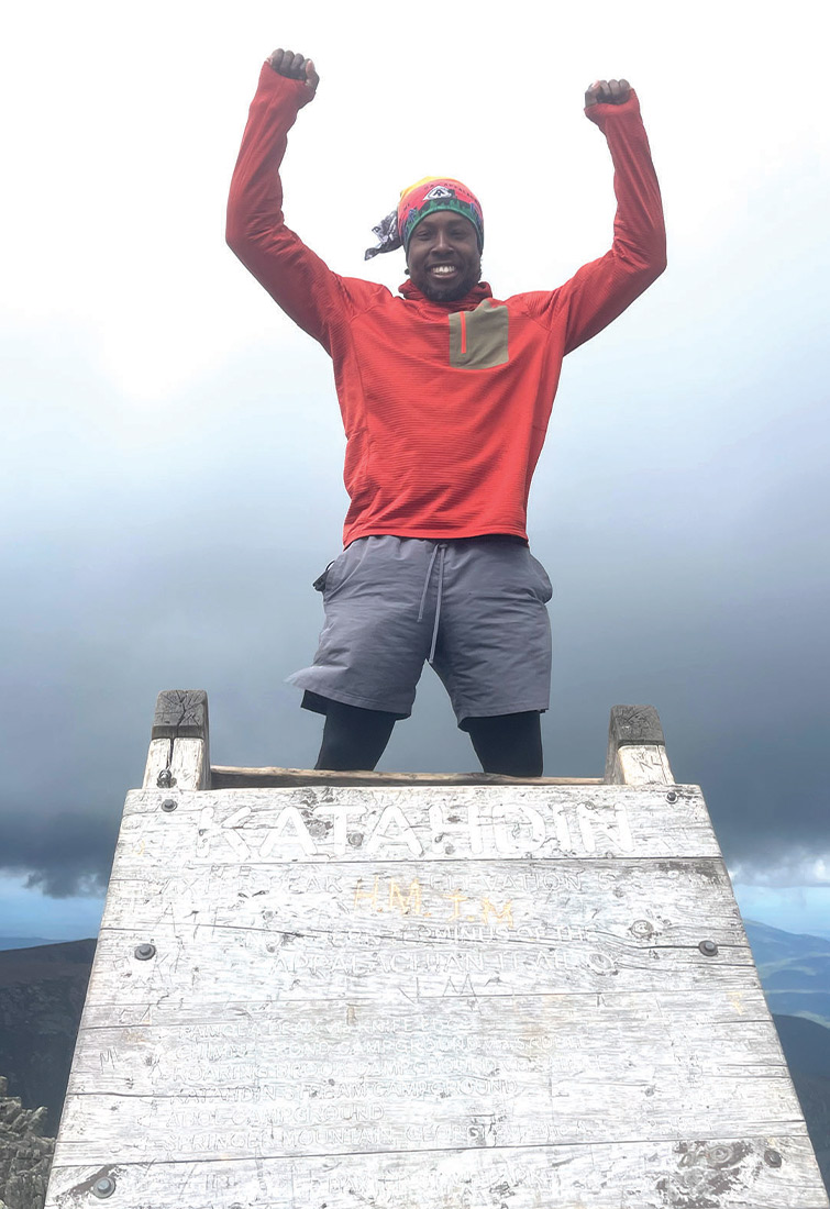 A smiling man stands on the weathered wooden Mount Katahdin summit sign with both arms raised. He wears a red long-sleeved shirt, grey shorts, and a colorful headband. The background is a bright, cloudy sky.