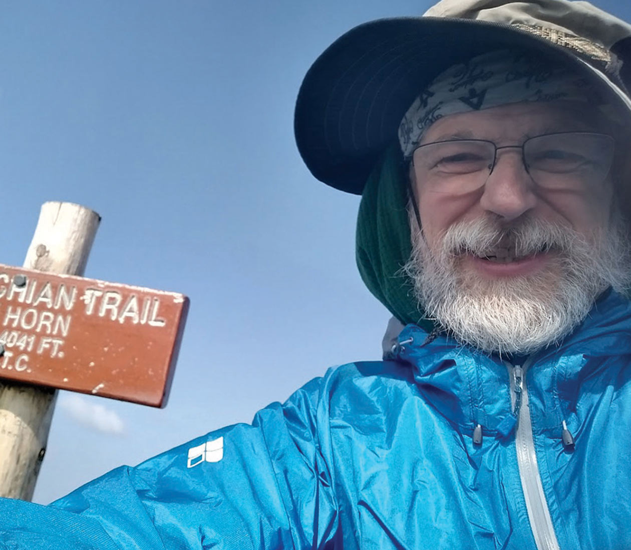 Selfie of smiling hiker George Jones with a white beard, wearing a blue jacket and hat. A wooden Appalachian Trail sign for The Horn at 4,041 feet stands beside him under a blue sky.