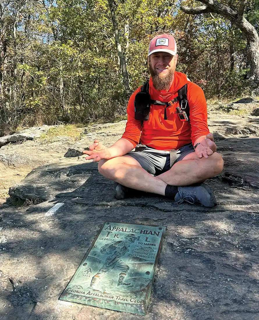 A man with a beard sits cross-legged on a large flat rock with his hands resting on his knees. He wears an orange shirt and a cap. A bronze Appalachian Trail plaque is in front of him on the rock. Sunlit trees are in the background.