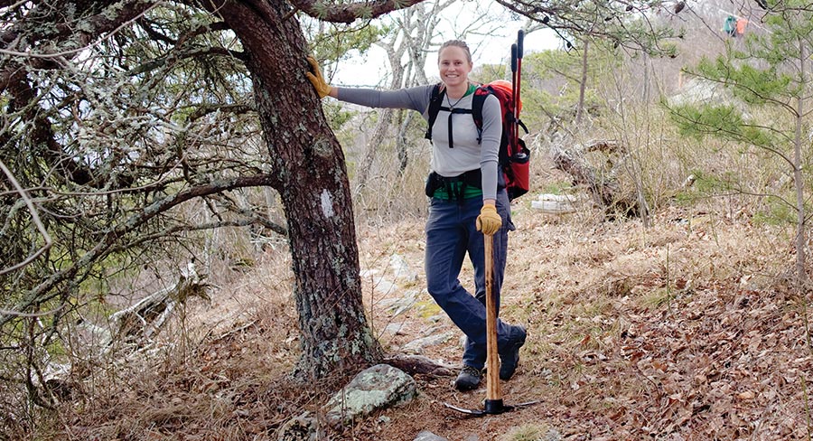A smiling woman with a backpack and gloves stands on a trail, leaning one hand against a tree with a white blaze. She holds a pickaxe upright. The surrounding forest floor is covered in dry leaves and low brush.