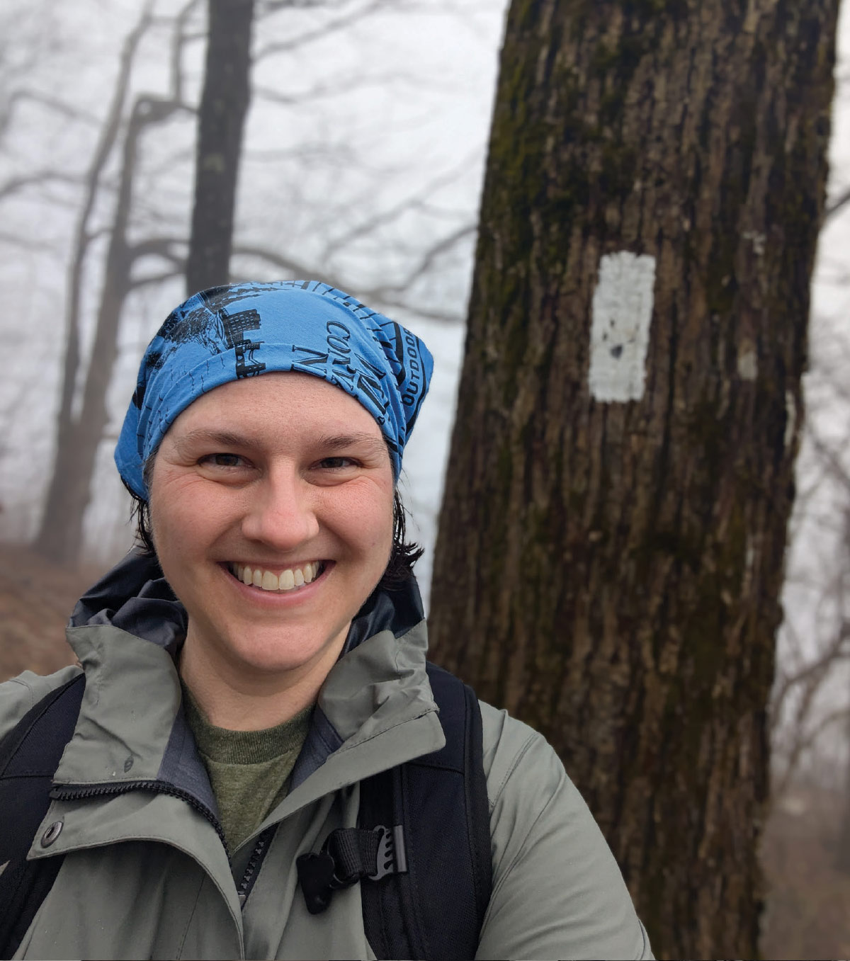 Monica Mogilewsky, a smiling woman standing at Blood Mountain, GA in a grey technical jacket and a vibrant blue patterned buff or bandana on her head