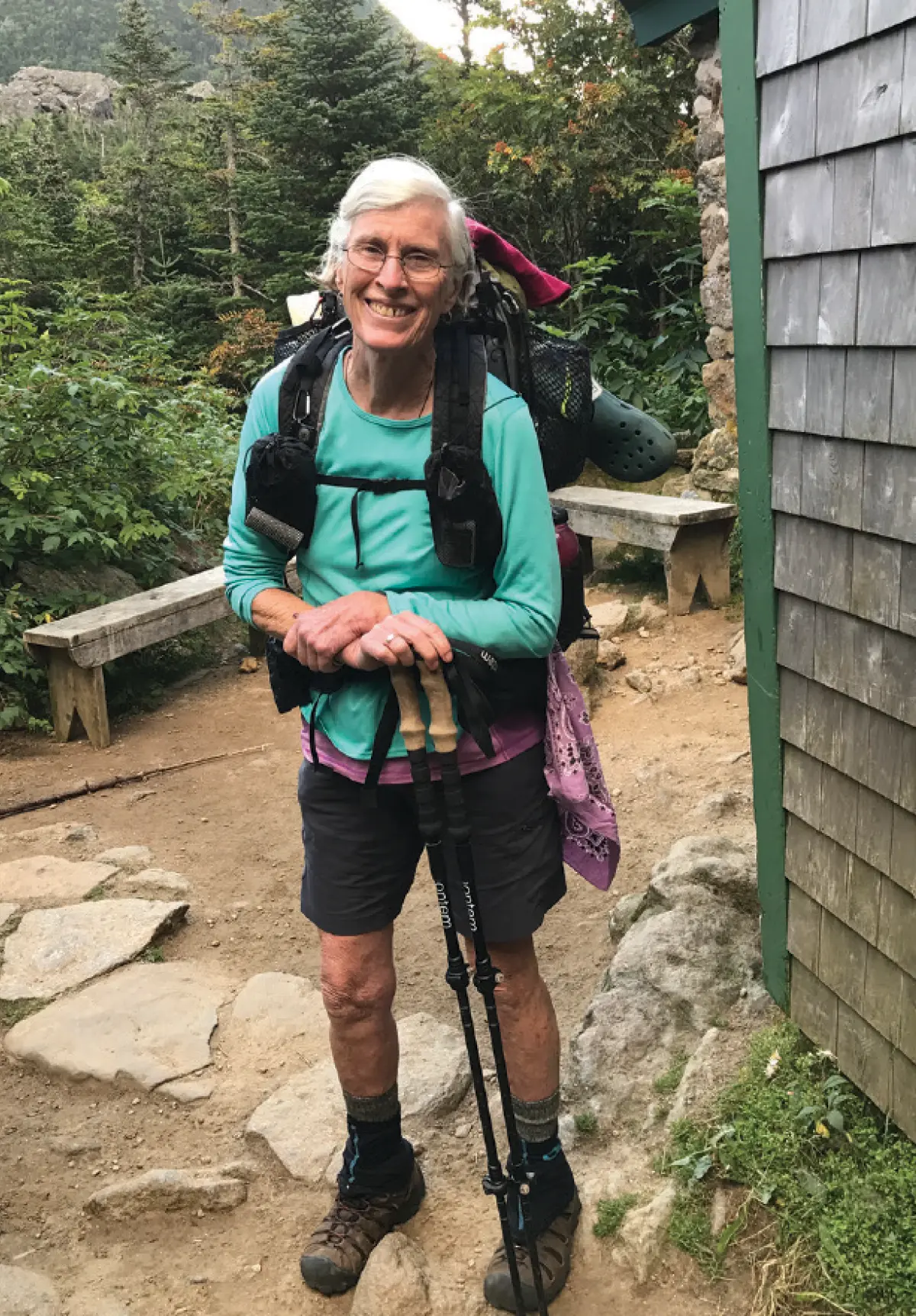 Mary Blanton, a smiling woman with short white hair standing on a rocky trail outfitted for a long-distance hike, leaning on two trekking poles