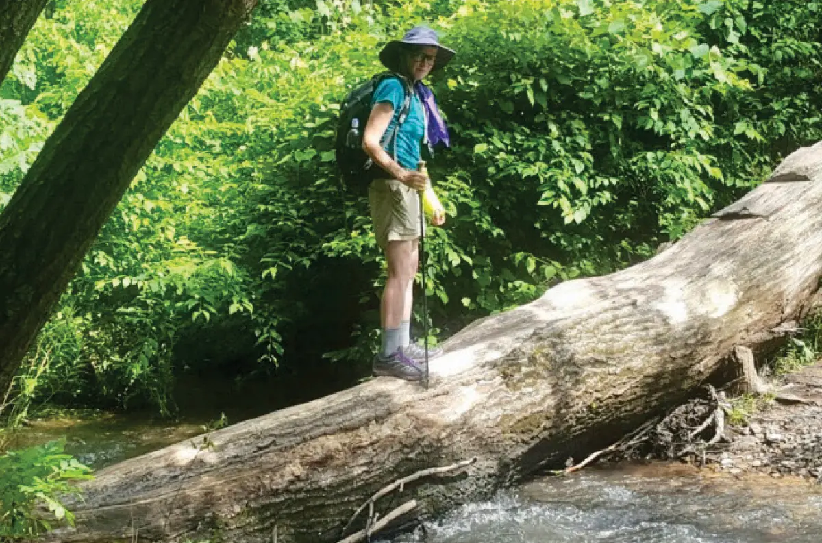 Charli Fulton, a smiling woman stands carefully atop a large, fallen tree trunk as she is in casual hiking attire