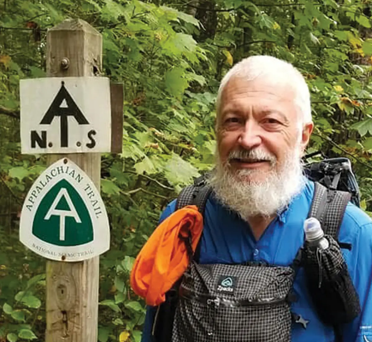 R. Dudley, a smiling man with a full white beard standing next to a wooden trail marker on the Appalachian Trail in casual hiking attire