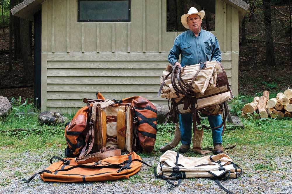 A man in a cowboy hat holds up heavy canvas pack gear next to large orange panniers outside a wooden trail shelter.