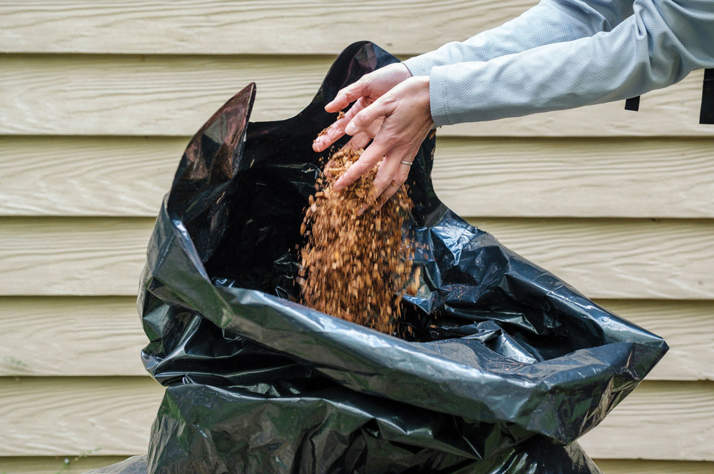 Close-up of hands pouring brown wood shavings or mulch out of a large, heavy-duty black plastic trash bag.