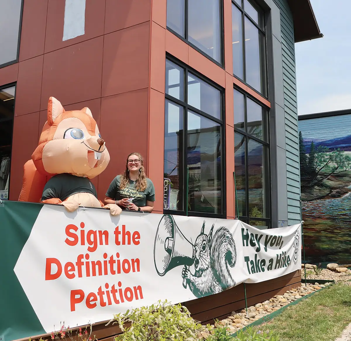 A smiling woman stands next to a large inflatable squirrel mascot behind a banner reading "Sign the Definition Petition" and "Hey you, Take a Hike" outside a modern building.
