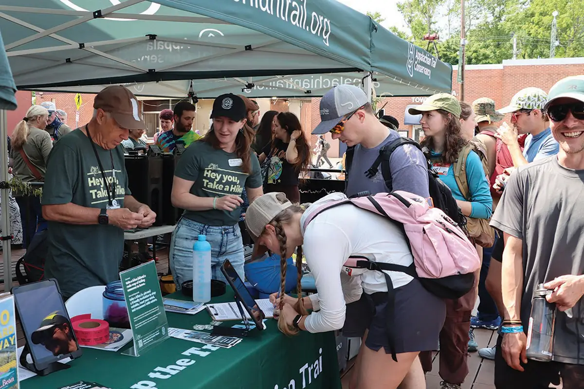 A woman with a pink backpack signs a document at an outdoor Appalachian Trail Conservancy booth. Staff wearing "Hey you, Take a Hike" t-shirts assist visitors under a green canopy tent.