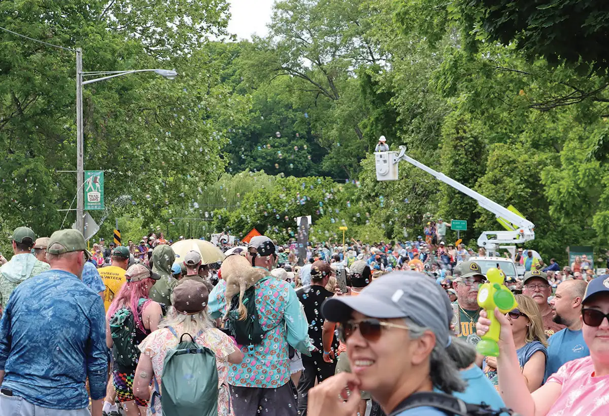 A large, festive crowd walks down a tree-lined street filled with hundreds of floating bubbles, with a bucket truck arm extended in the background.