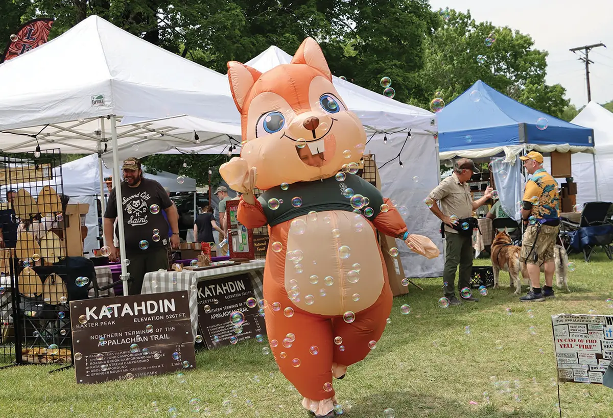An inflatable squirrel mascot dances through floating bubbles at an outdoor festival featuring event tents and an Appalachian Trail Katahdin sign.