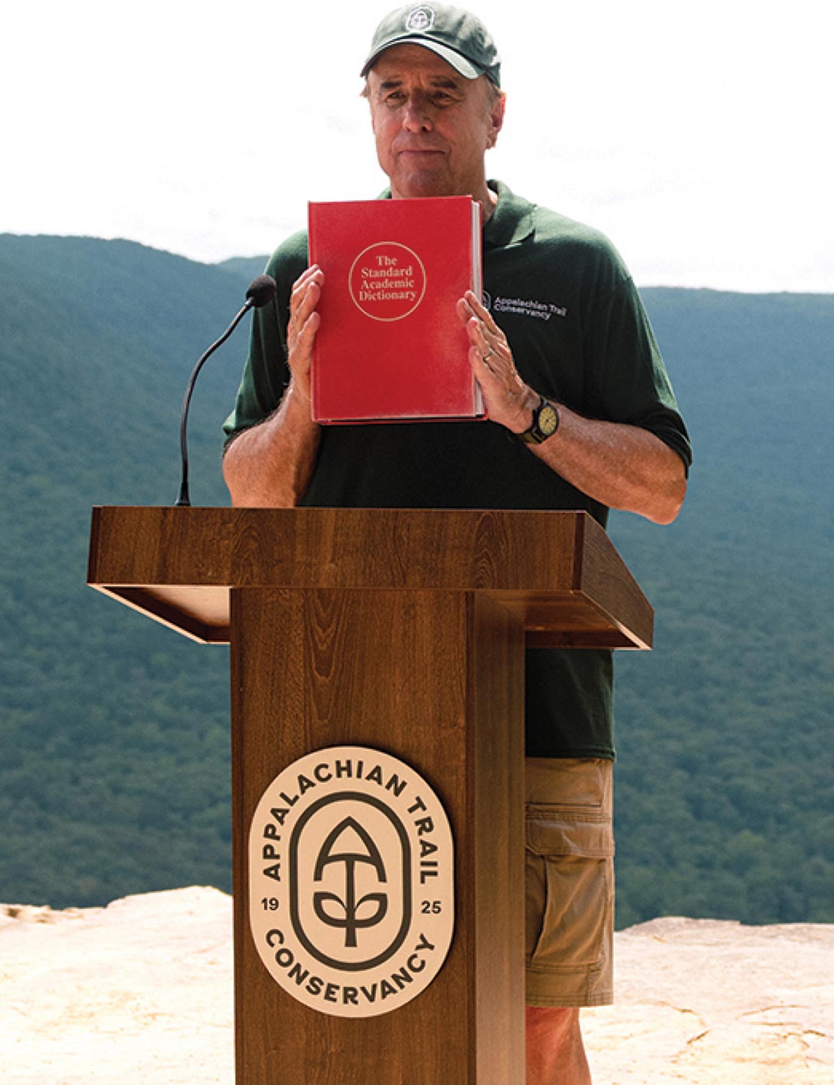 A man in a green Appalachian Trail Conservancy polo and cap stands at a logo podium, holding up a red book titled "The Standard Academic Dictionary" against a backdrop of forested hills.