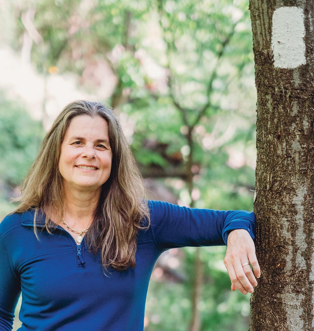Smiling woman in a blue pullover leaning against a tree trunk with a white trail blaze, surrounded by soft-focus green foliage.