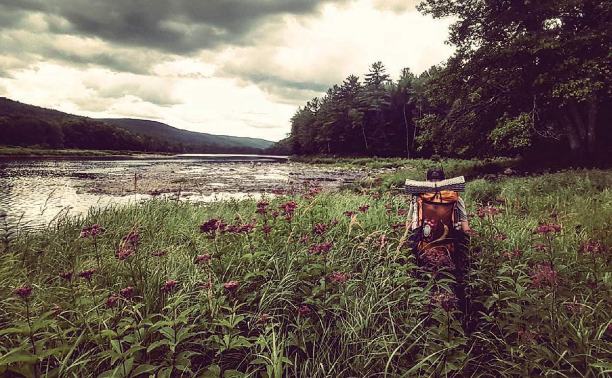 A backpacker walks through tall grass and pink wildflowers along a wide river bank under a dramatic, cloudy sky.