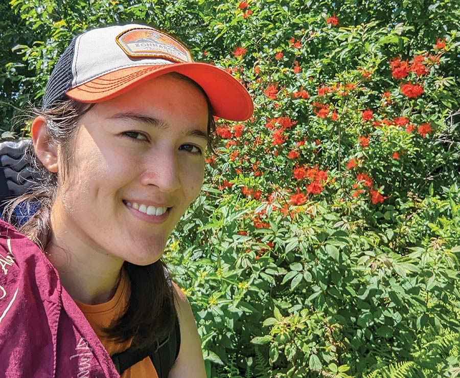 A smiling woman wearing a baseball cap takes a selfie outdoors in front of a green bush filled with bright orange flowers.