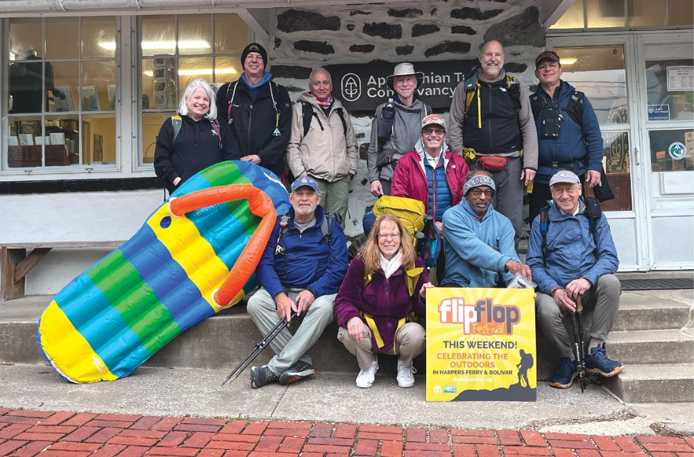 Eleven hikers pose outside an Appalachian Trail Conservancy building beside a giant inflatable flip-flop and a Flip Flop Festival sign.