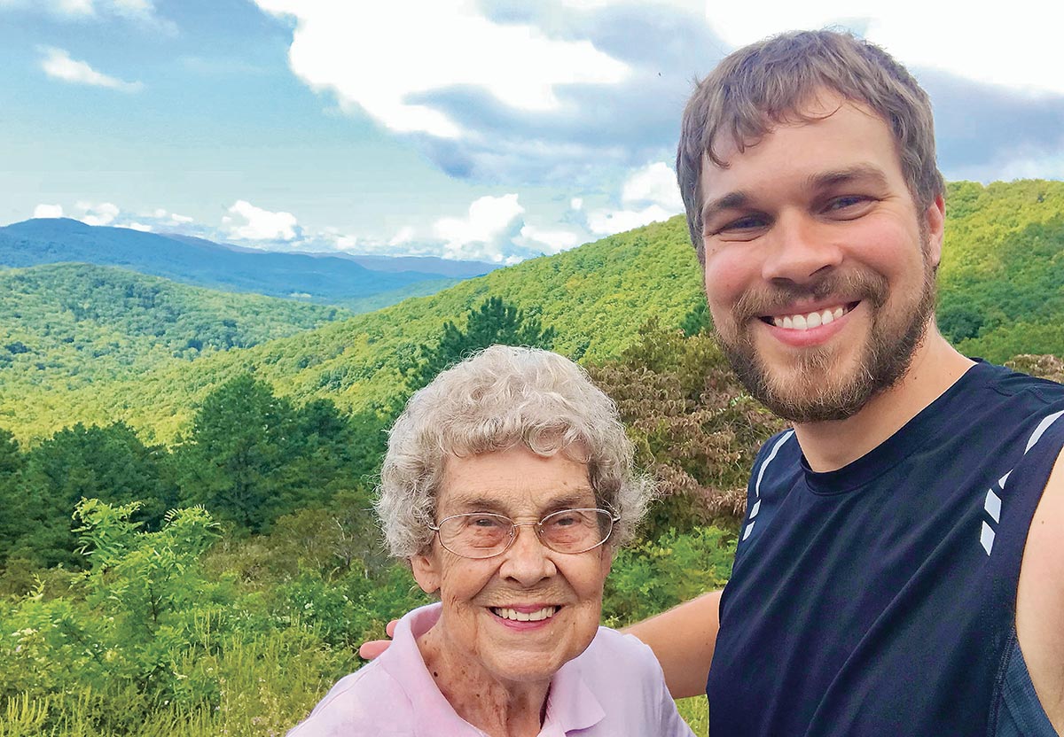 A man and an older woman smile for a selfie in front of rolling green mountains.