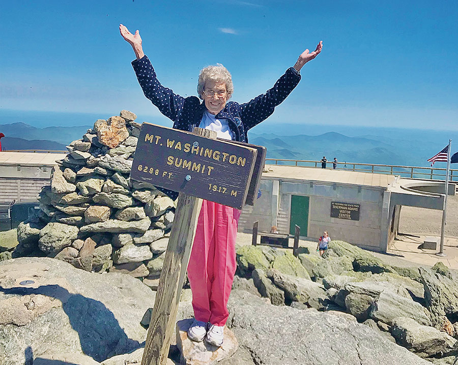 An older woman raises her arms joyfully beside the Mount Washington summit sign.
