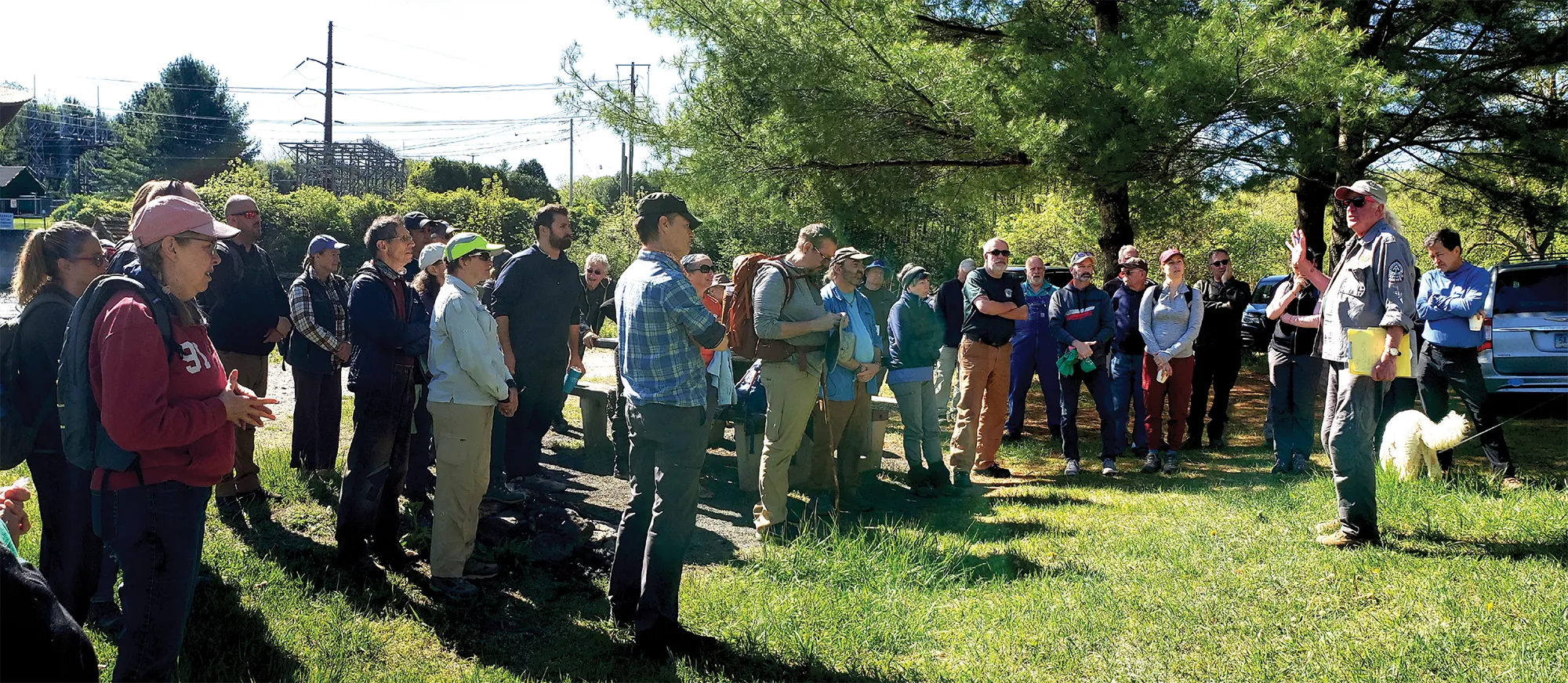A group of volunteers listens to a speaker during an outdoor gathering beneath trees.