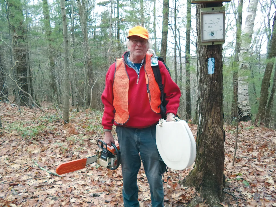 Trail volunteer in an orange safety vest and cap holds a chainsaw and protective helmet beside a marked tree.