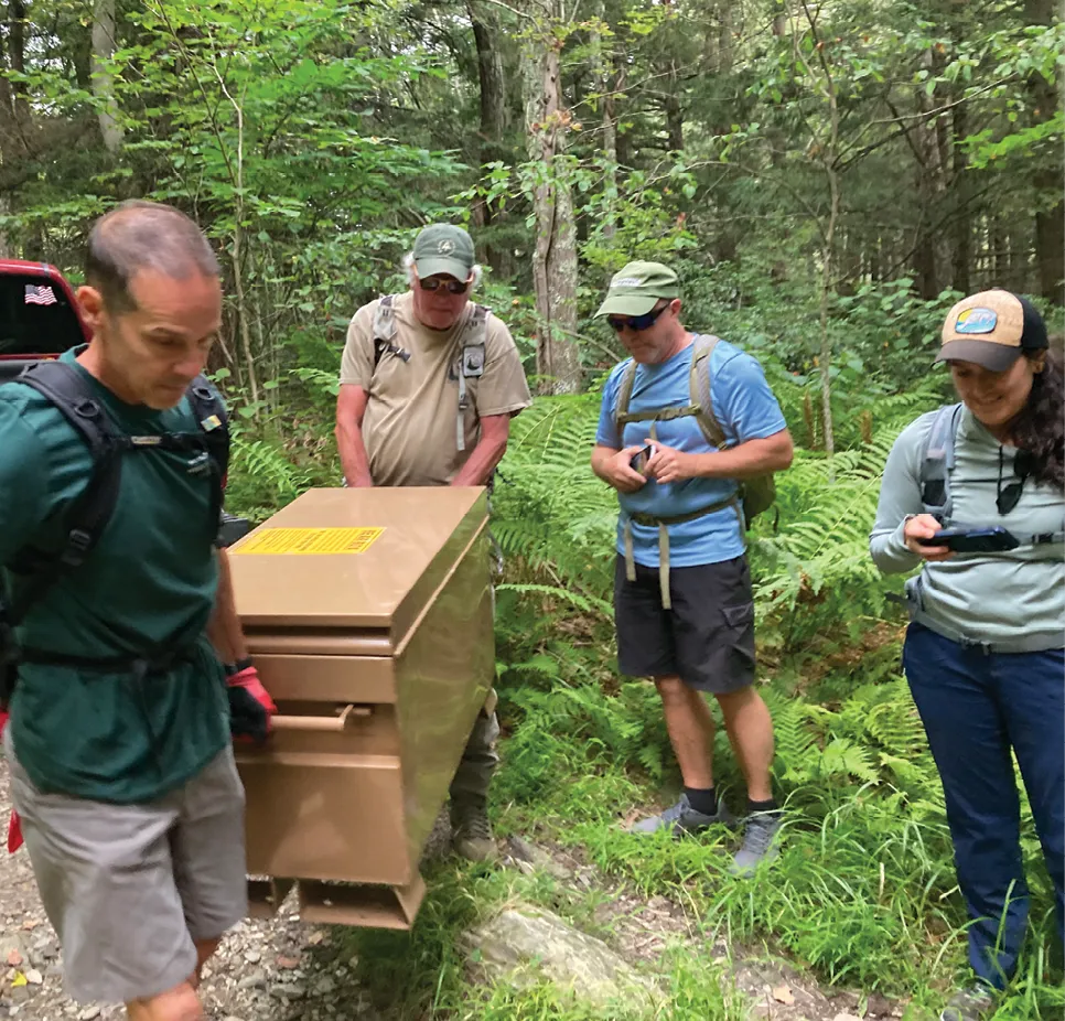 Volunteers carry a large wooden trail structure through a fern-filled forest while hikers stand nearby.